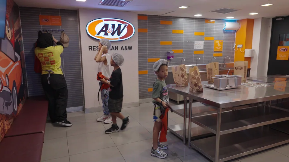 孩子戴著髮網，站在 A&W 餐飲體驗的工作台旁。