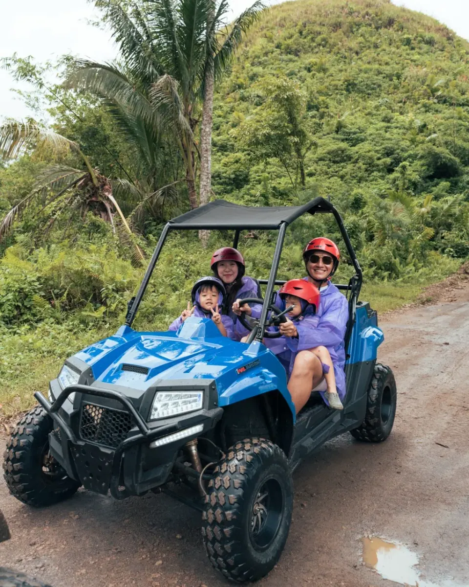 一家四口穿紫色雨衣、戴安全帽坐在藍色 buggy 上,後方是山丘與椰子樹。