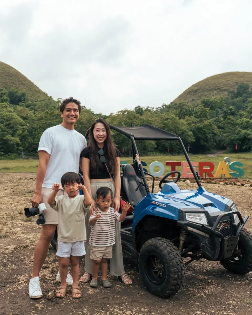 一家四口站在藍色 buggy 旁合照,後方是場地招牌與綠色丘陵。