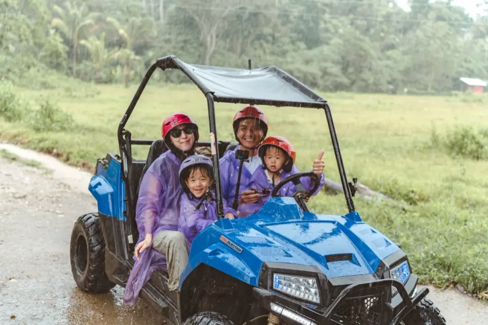 雨中一家四口穿雨衣坐在藍色 buggy 裡,車頂與道路濕亮。
