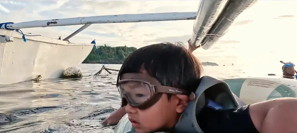 戴面鏡的孩子在船邊水面,旁邊有成人手臂扶著。