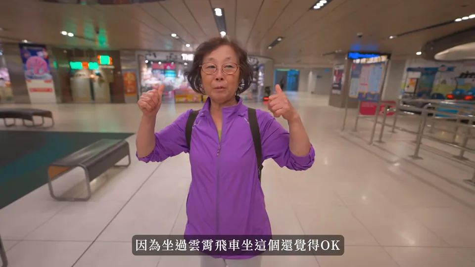 阿婆穿著紫色上衣,在航廈內面向鏡頭,雙手比出拇指。